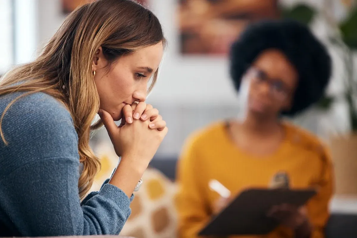 A woman sits deep in thought during a therapy session, with a therapist listening attentively in the background.