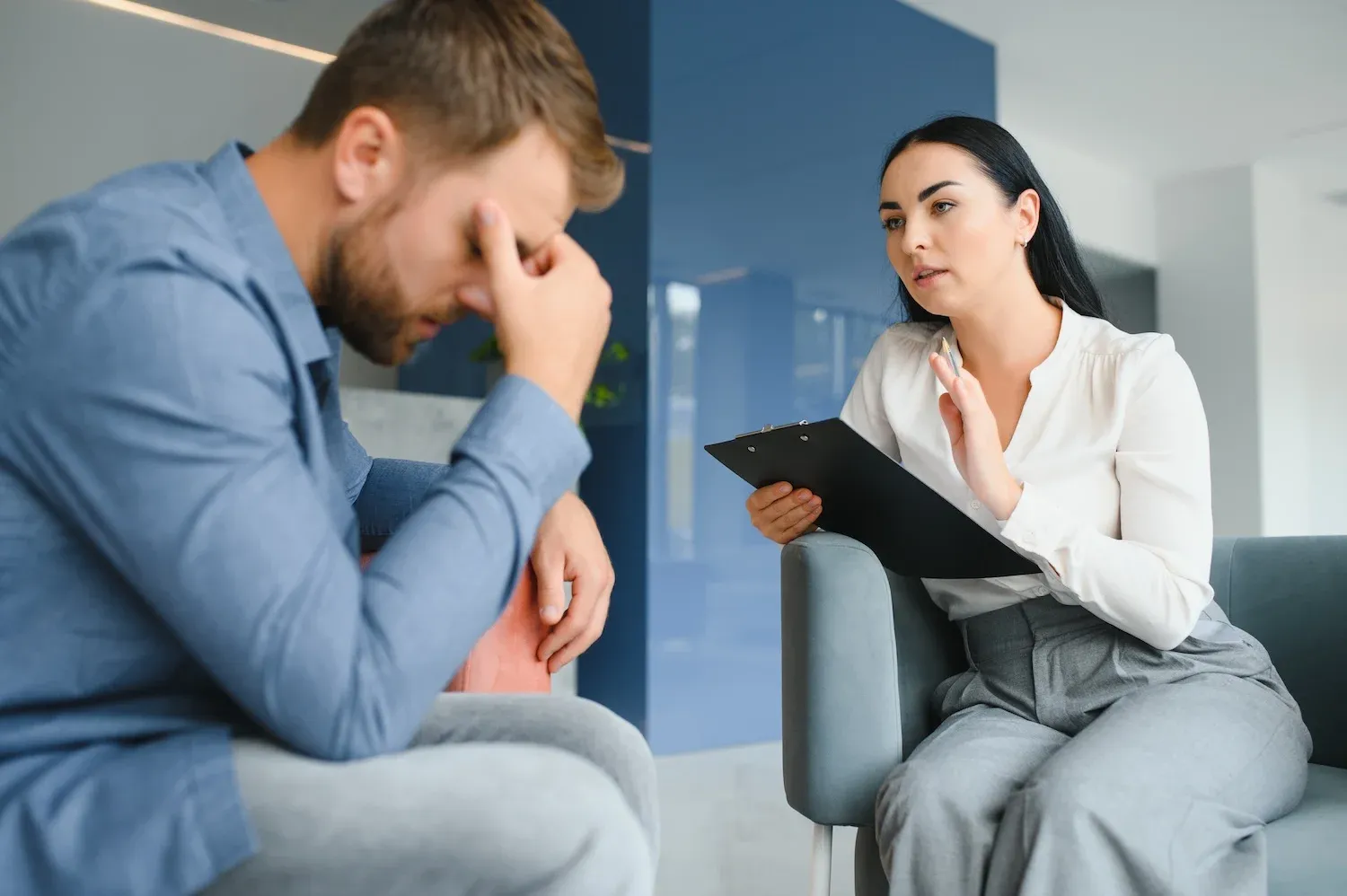 Man holding his head in distress while talking with a therapist holding a clipboard.