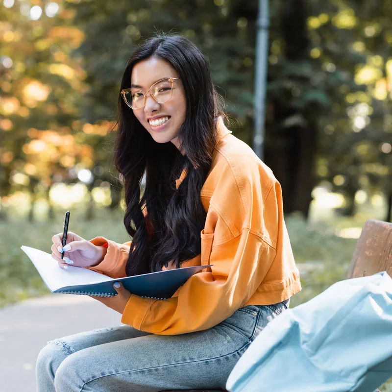 a young woman with glasses pauses writing in her notebook to look up and smile at the camera
