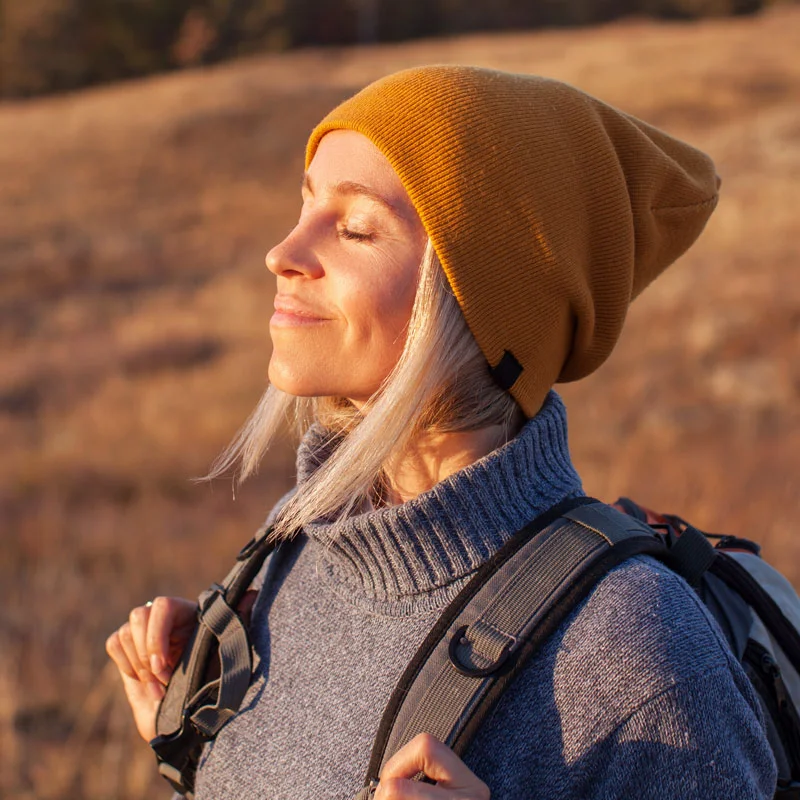 a smiling woman with a backpack and beanie turns to the sun with eyes closed and smiles
