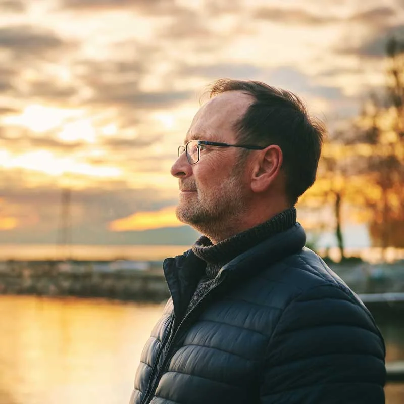 a pensive older man with glasses looks off into the sunset