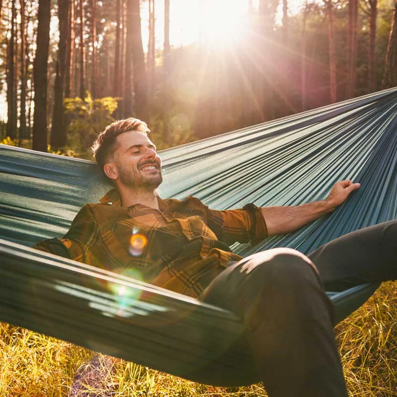 a smiling young man relaxes in a hammock set up in a wooded area