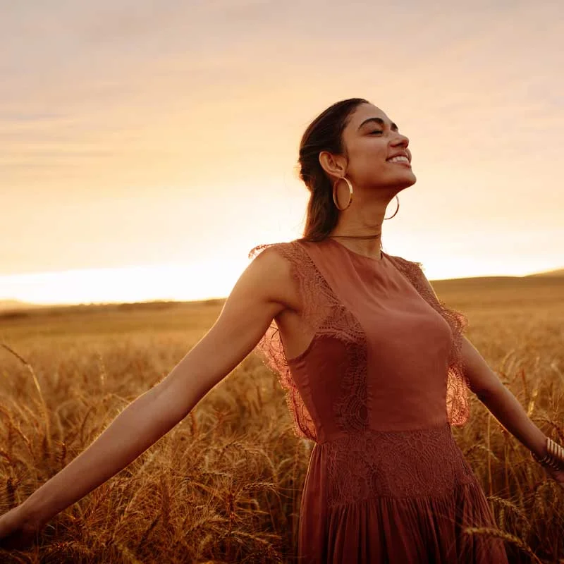 a smiling woman stands with arms outstretched in a wheat field at sunset