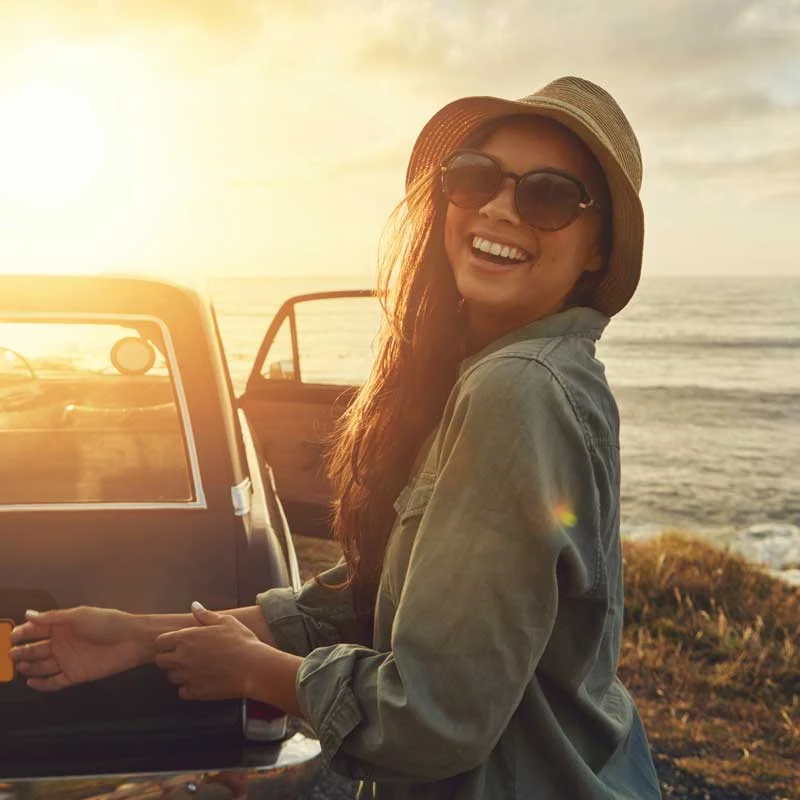 a young woman in big sunglasses and a big hat smiles at the camera on the beach
