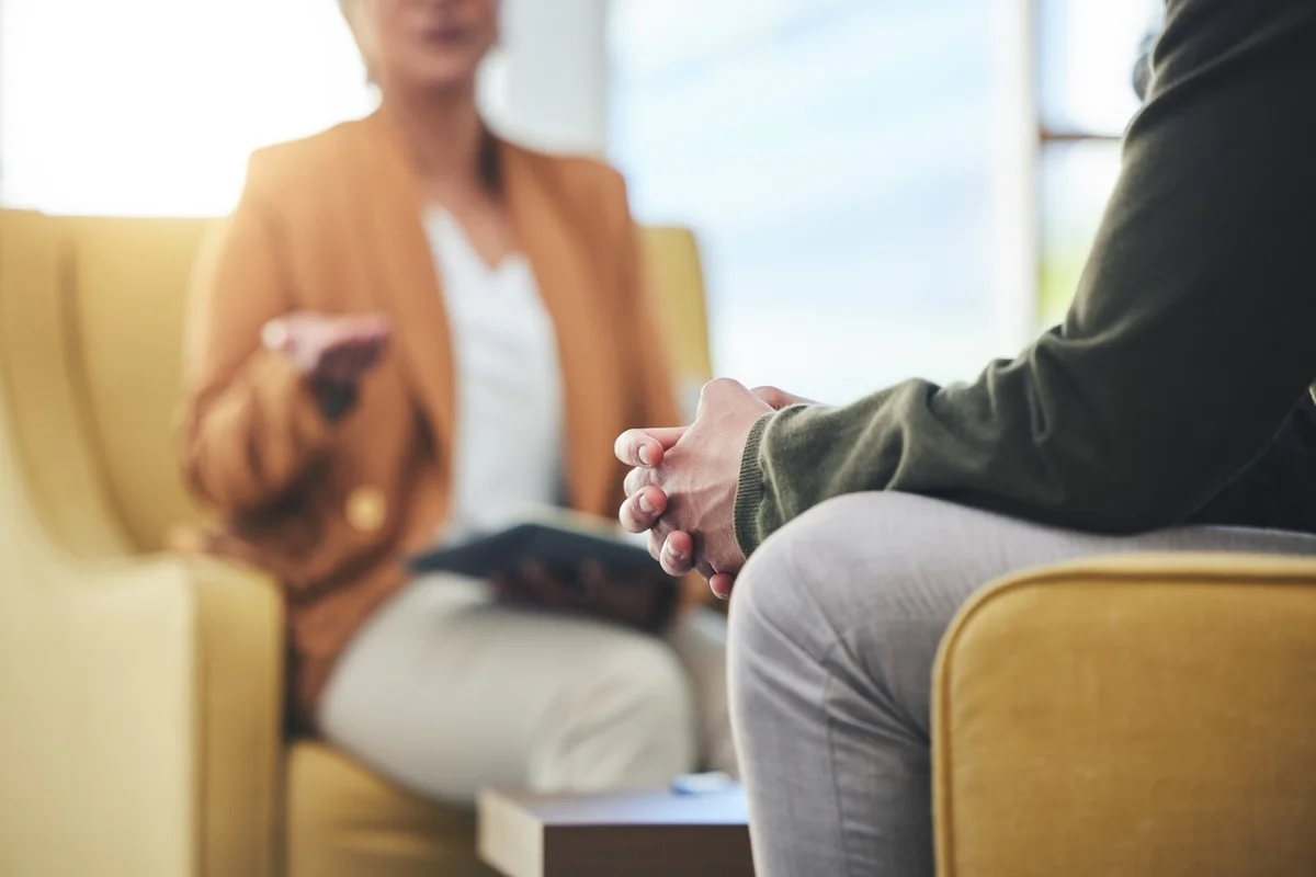A person sits with hands clasped during a one-on-one therapy session as a counselor gestures while speaking.