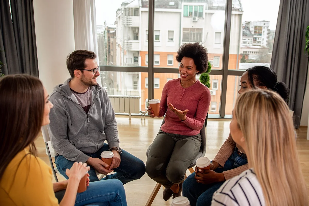 A diverse group of people sit together in a bright room, smiling and talking while holding coffee cups during a group therapy meeting.