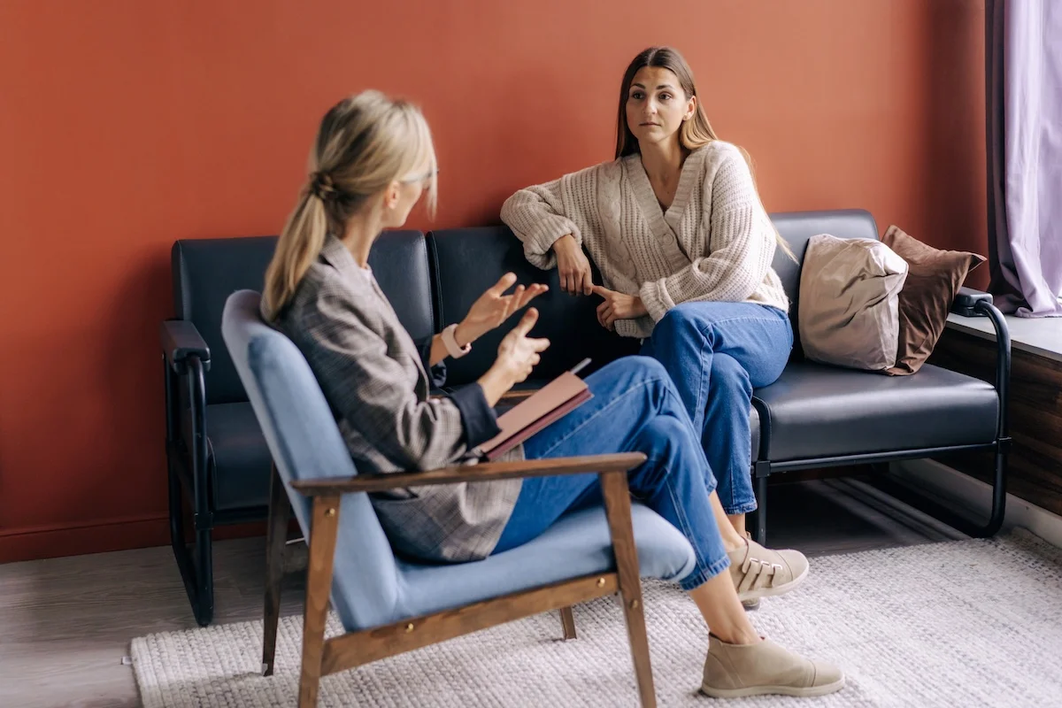 A therapist speaks with a woman during a counseling session in a cozy room with a warm-toned wall.
